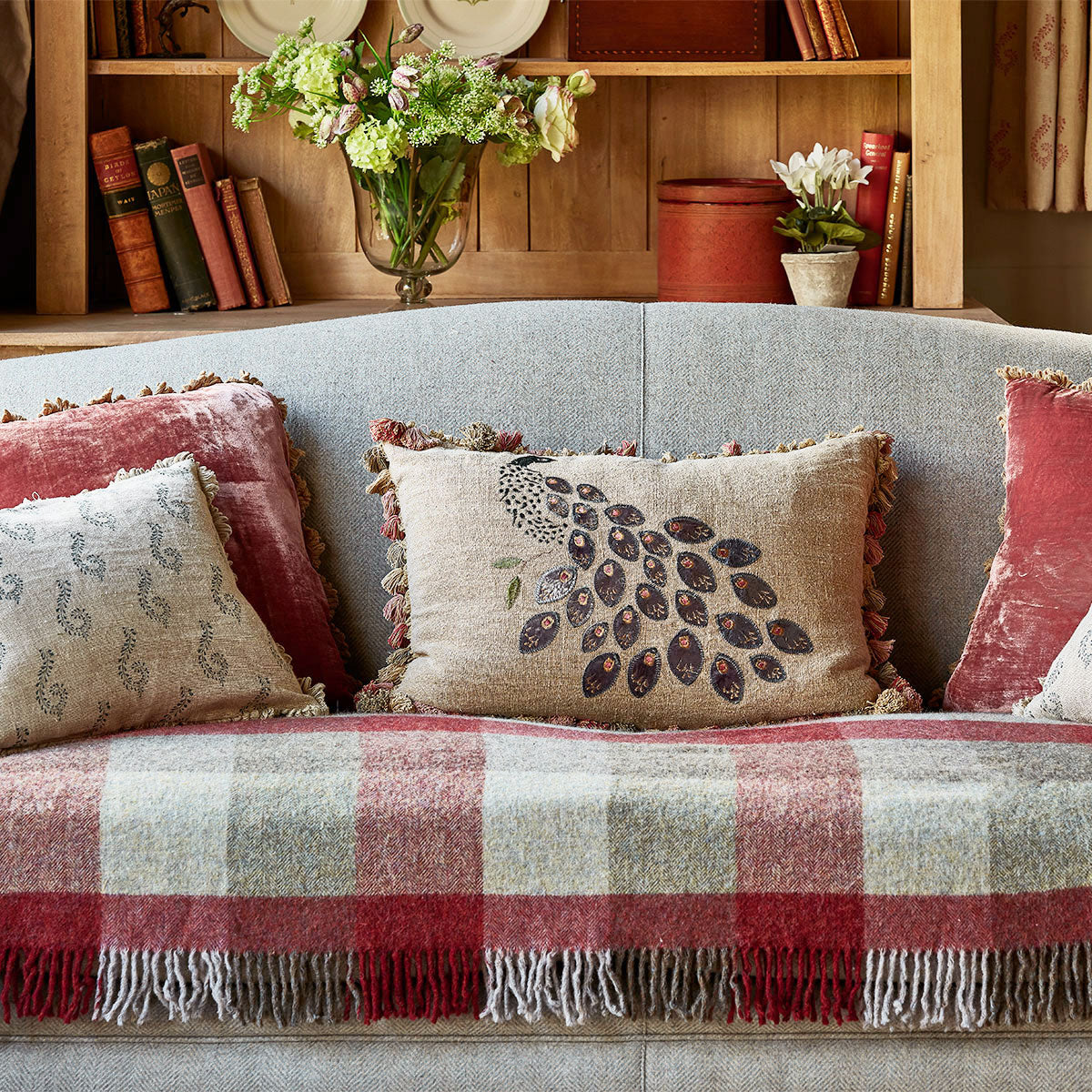 Peacock cushions on a sofa with a red checked throw and bookshelves