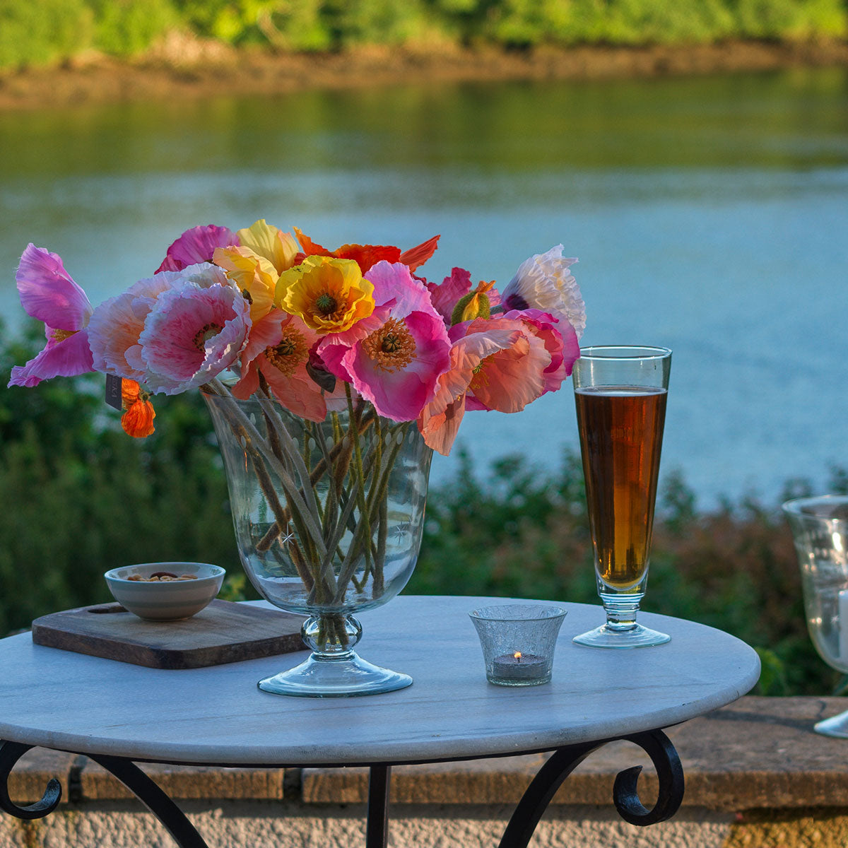 Hand-blown beer glass on an outdoor table with flowers beside water