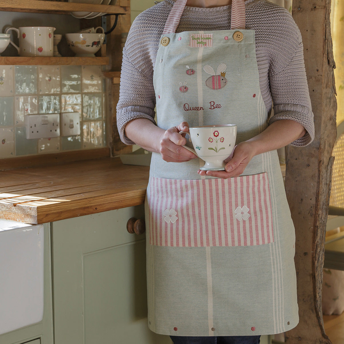 Woman in the embroidered Queen Bee apron holding a mug in a kitchen