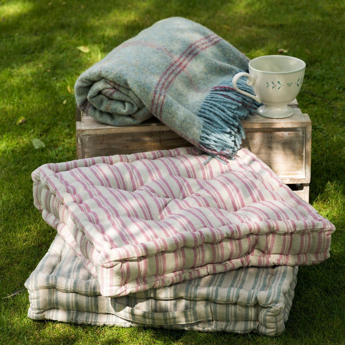 Pink and duck egg box cushions stacked on grass with a folded throw