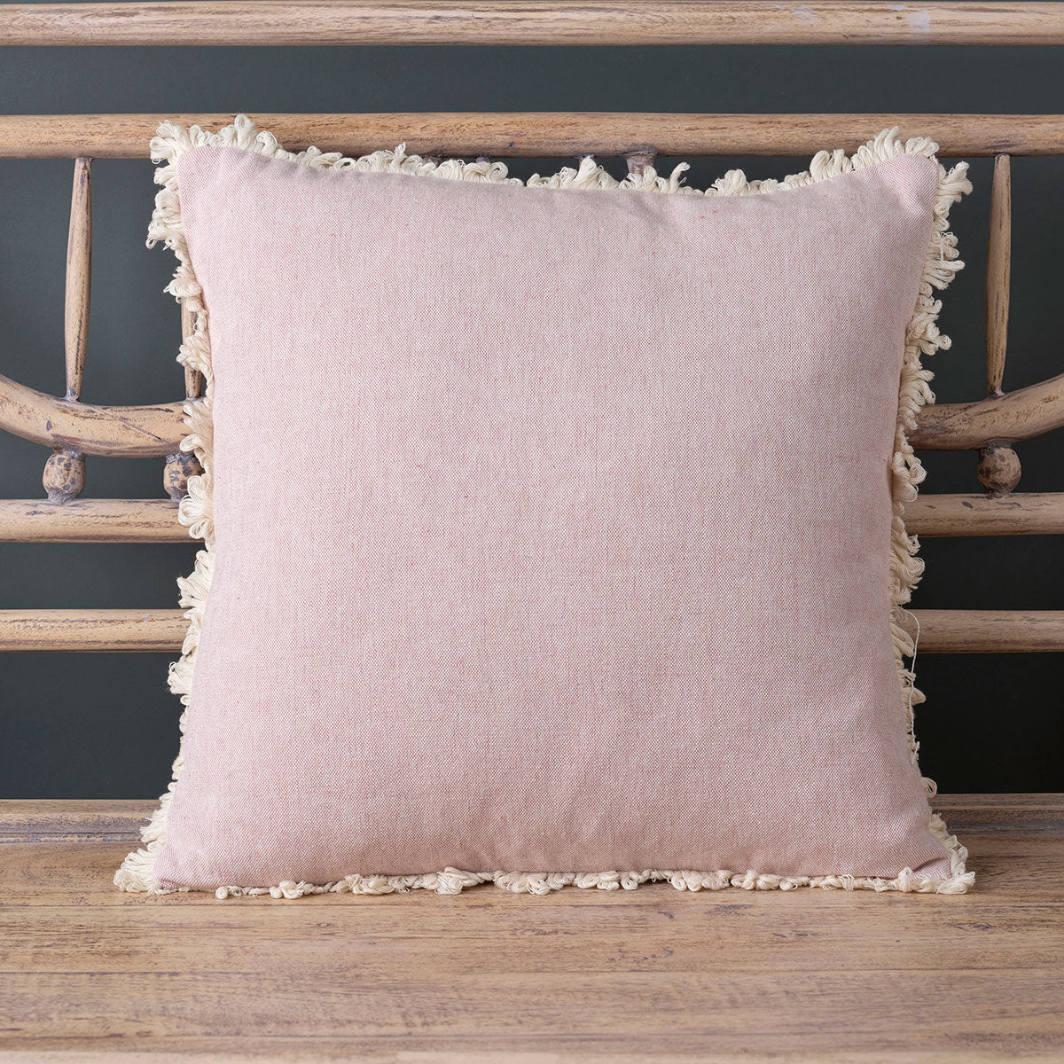 Pale rose plain cotton cushion with a soft fringed edge on a wooden bench