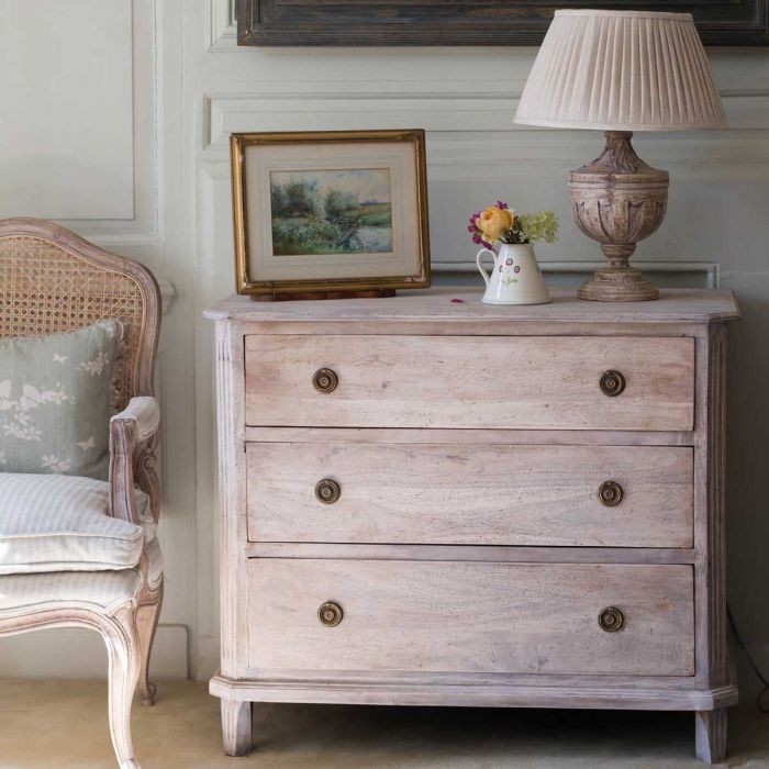 Classic chest of drawers in a panelled room beneath a lamp and painting