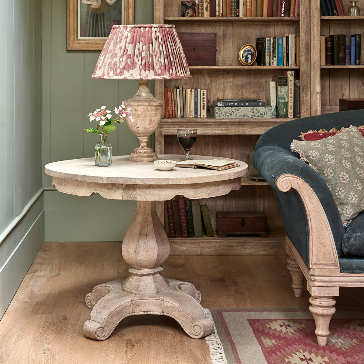 Round pedestal table with a pleated lampshade and flowers in a book-lined room